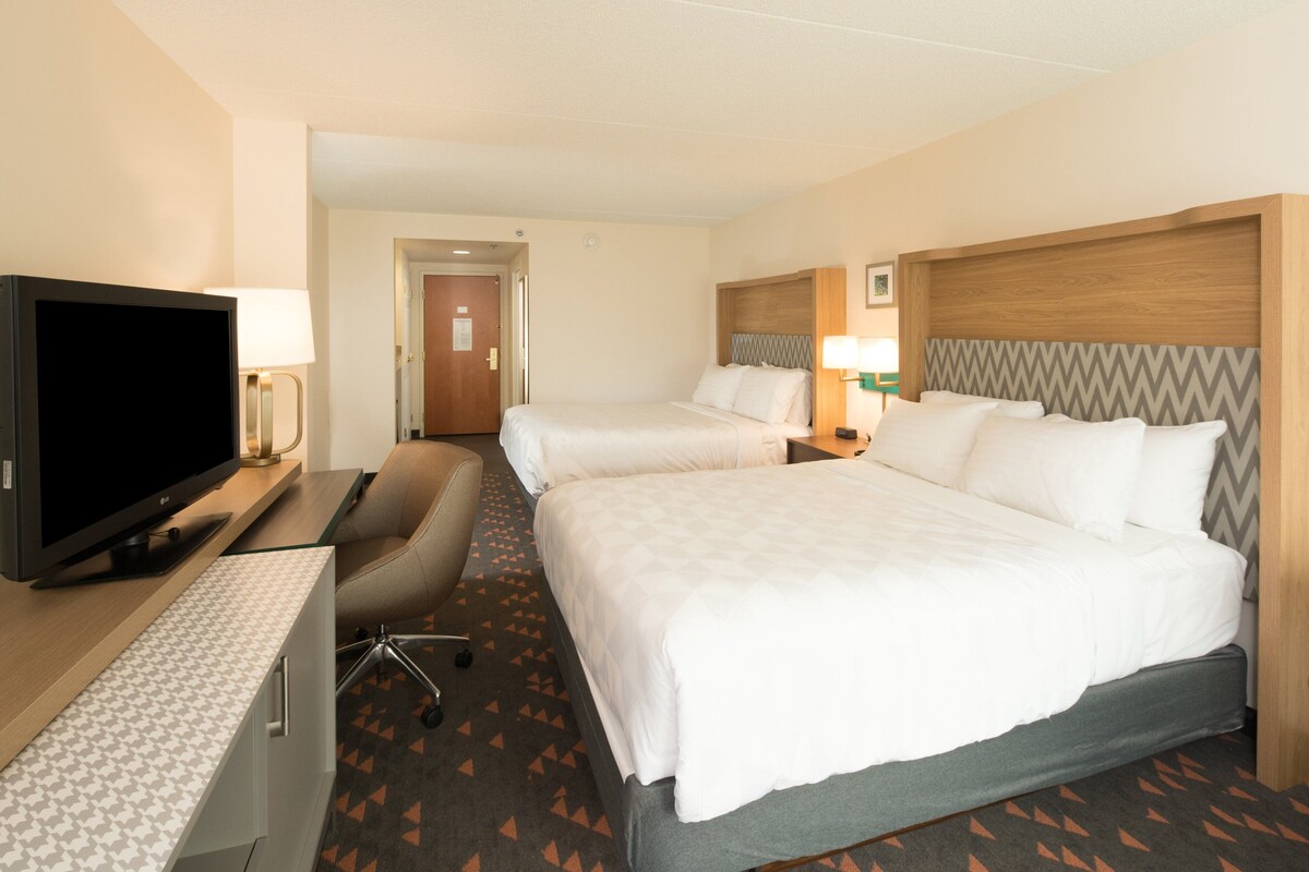 A hotel room with two beds, a desk with a TV, a chair, and a lamp.