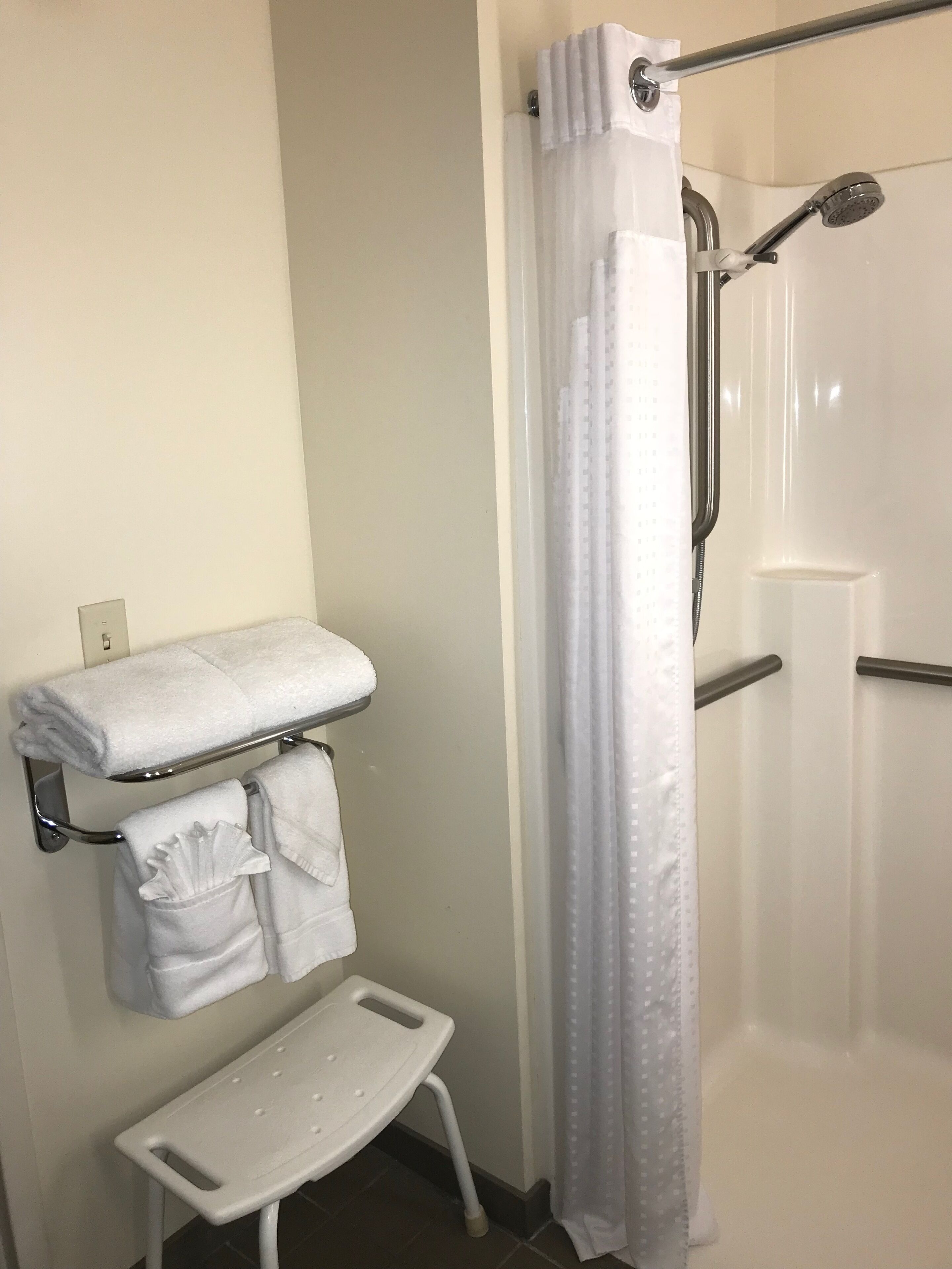 A bathroom with a shower, a towel rack with folded towels, and a small white stool.