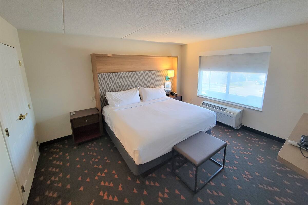 A hotel room with a bed, bedside table, lamp, window, and a small bench.