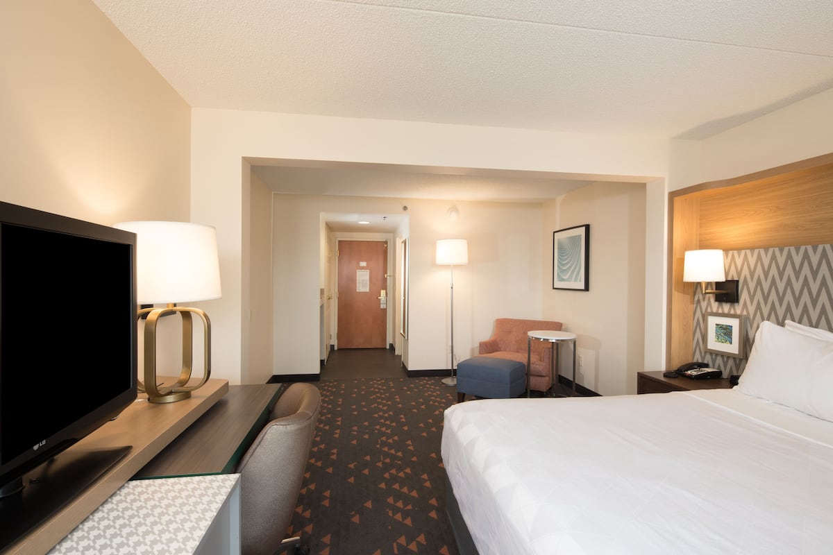 A hotel room with a bed, a TV, a desk with a lamp, a chair, a small table, and a nightstand with a lamp.