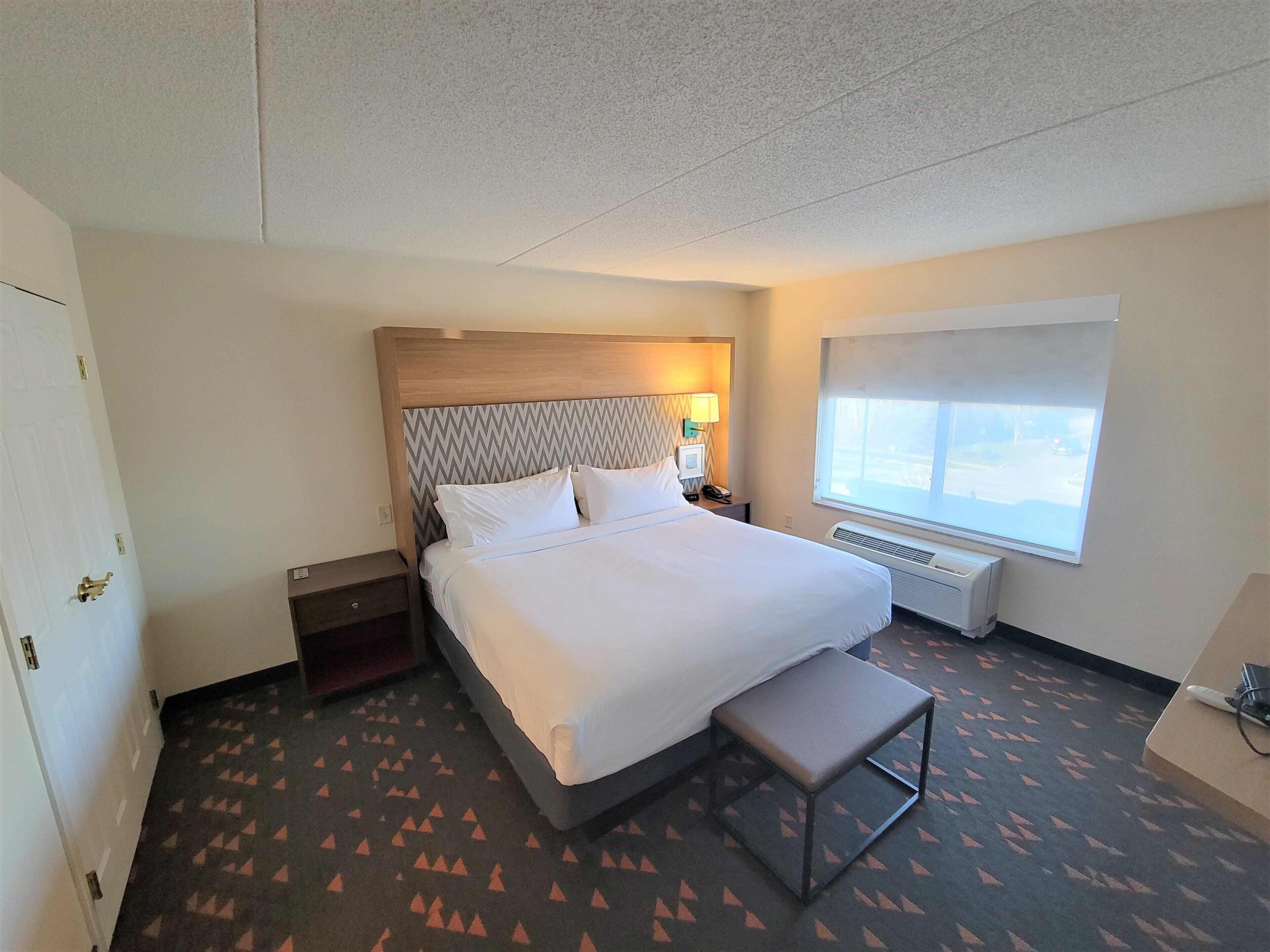 A hotel room with a bed, bedside table, nightstand, window, and a bench.