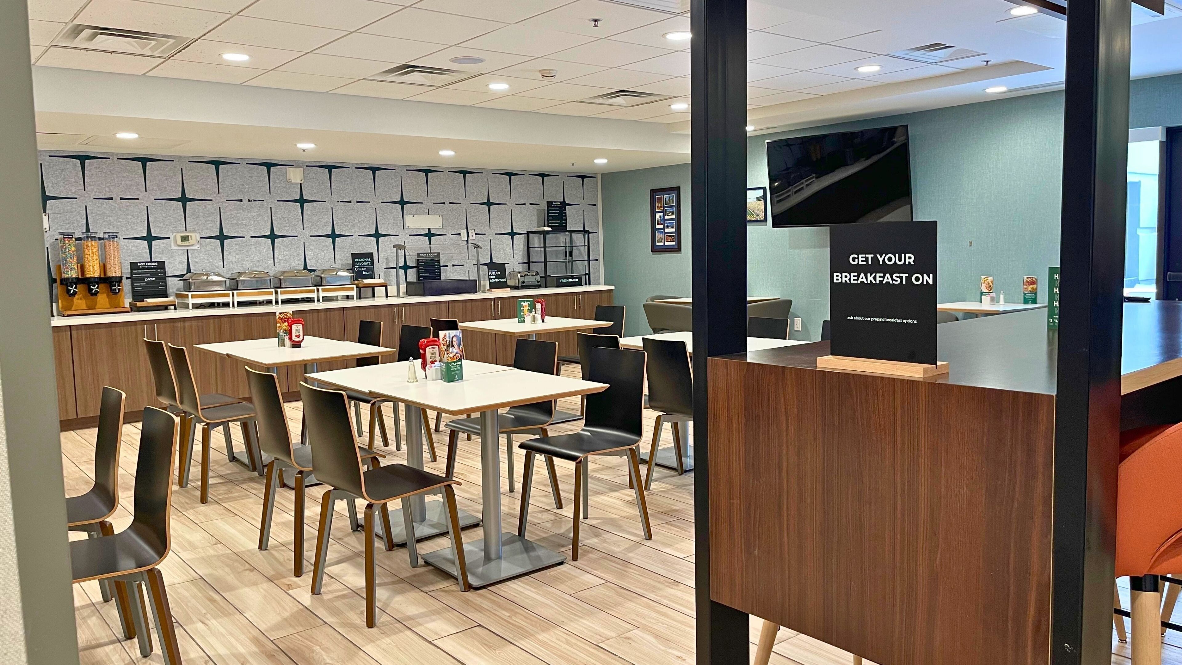 A modern breakfast area with tables and chairs, a counter with food dispensers, and a sign advertising breakfast options.