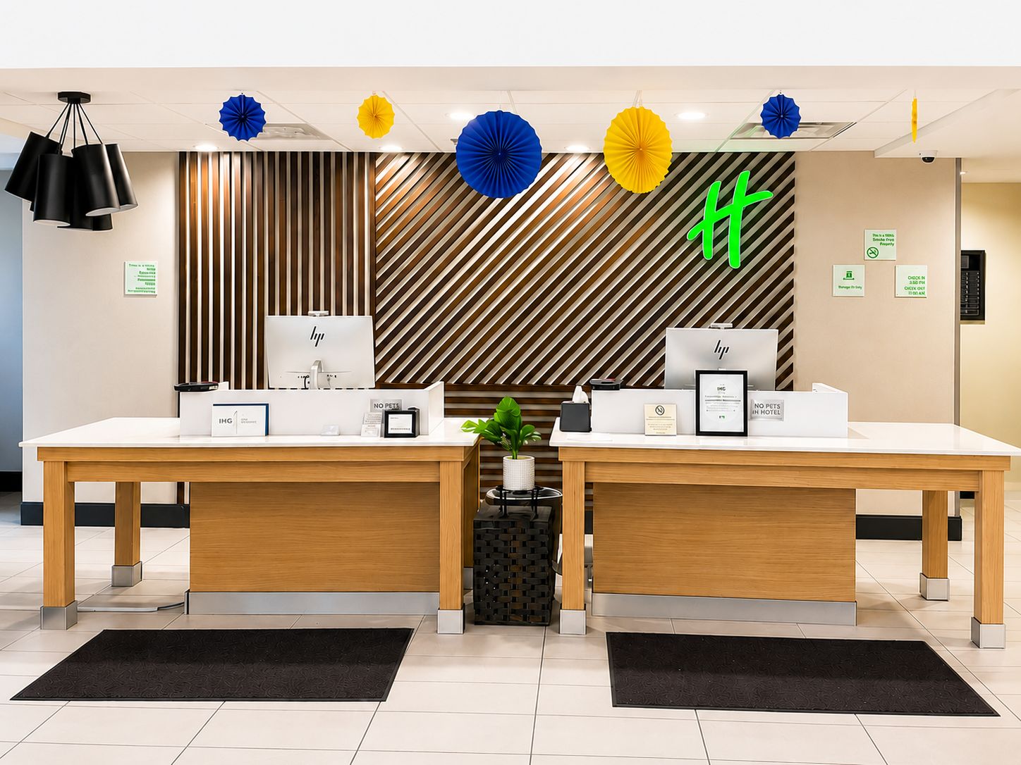 Un mostrador de recepción de hotel con dos estaciones de trabajo de ordenador, una planta y un panel decorativo con un diseño.