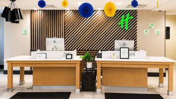 Un mostrador de recepción de hotel con dos estaciones de trabajo de ordenador, una planta y un panel decorativo con un diseño.