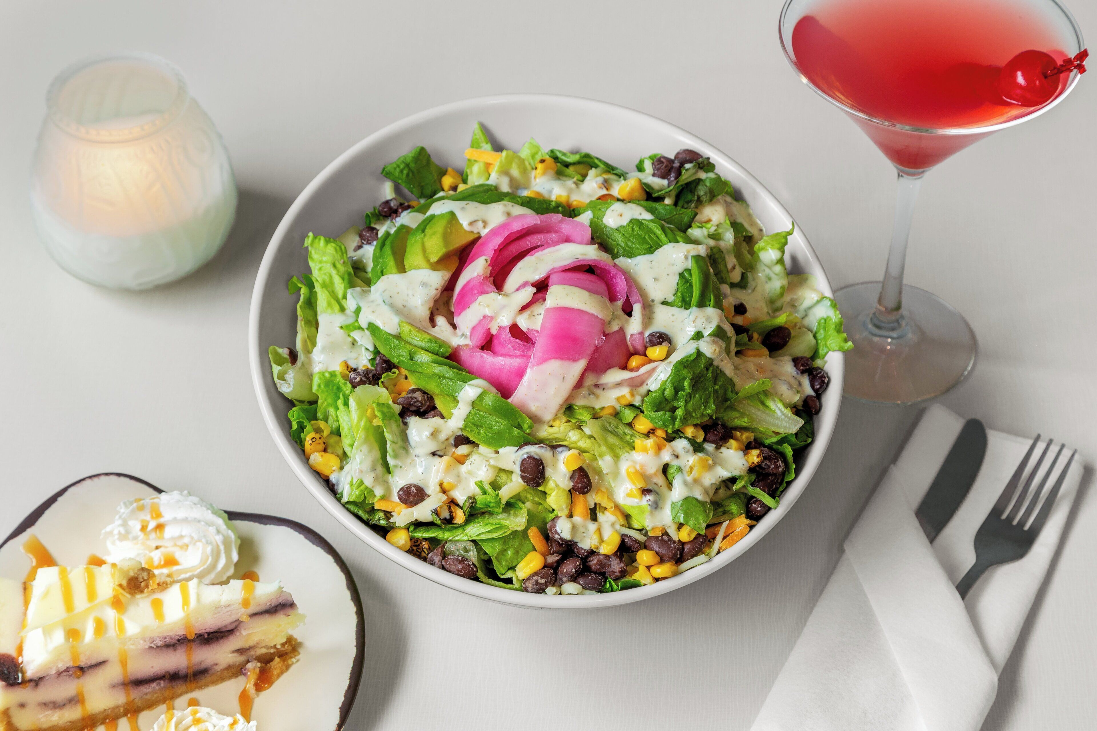Ensalada con diversos vegetales y legumbres, una porción de pastel de queso y un cóctel rojo a un lado.