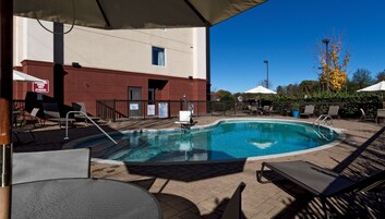 Una piscina con sillas de descanso y sombrillas, rodeada por un patio de ladrillo y un edificio de fachada roja.
