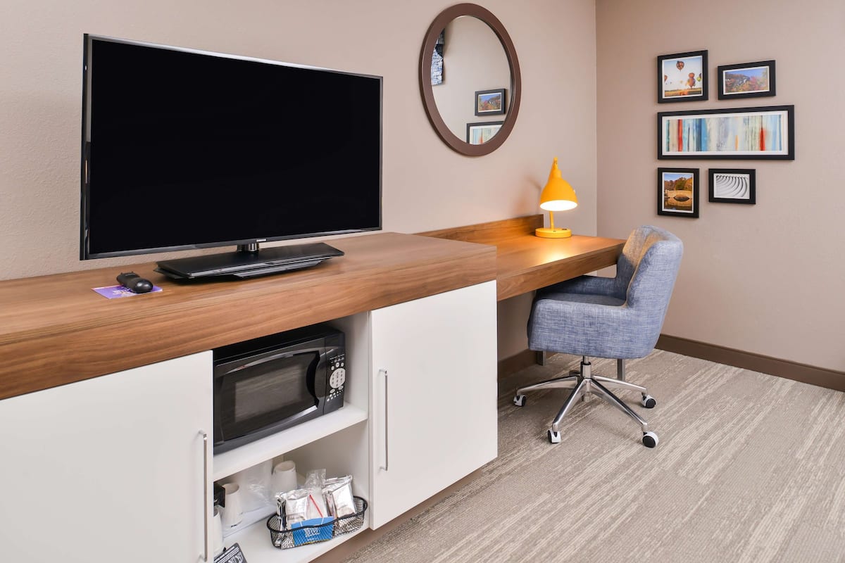A hotel room with a wooden desk, a flat-screen TV, a microwave, a chair, and a lamp.