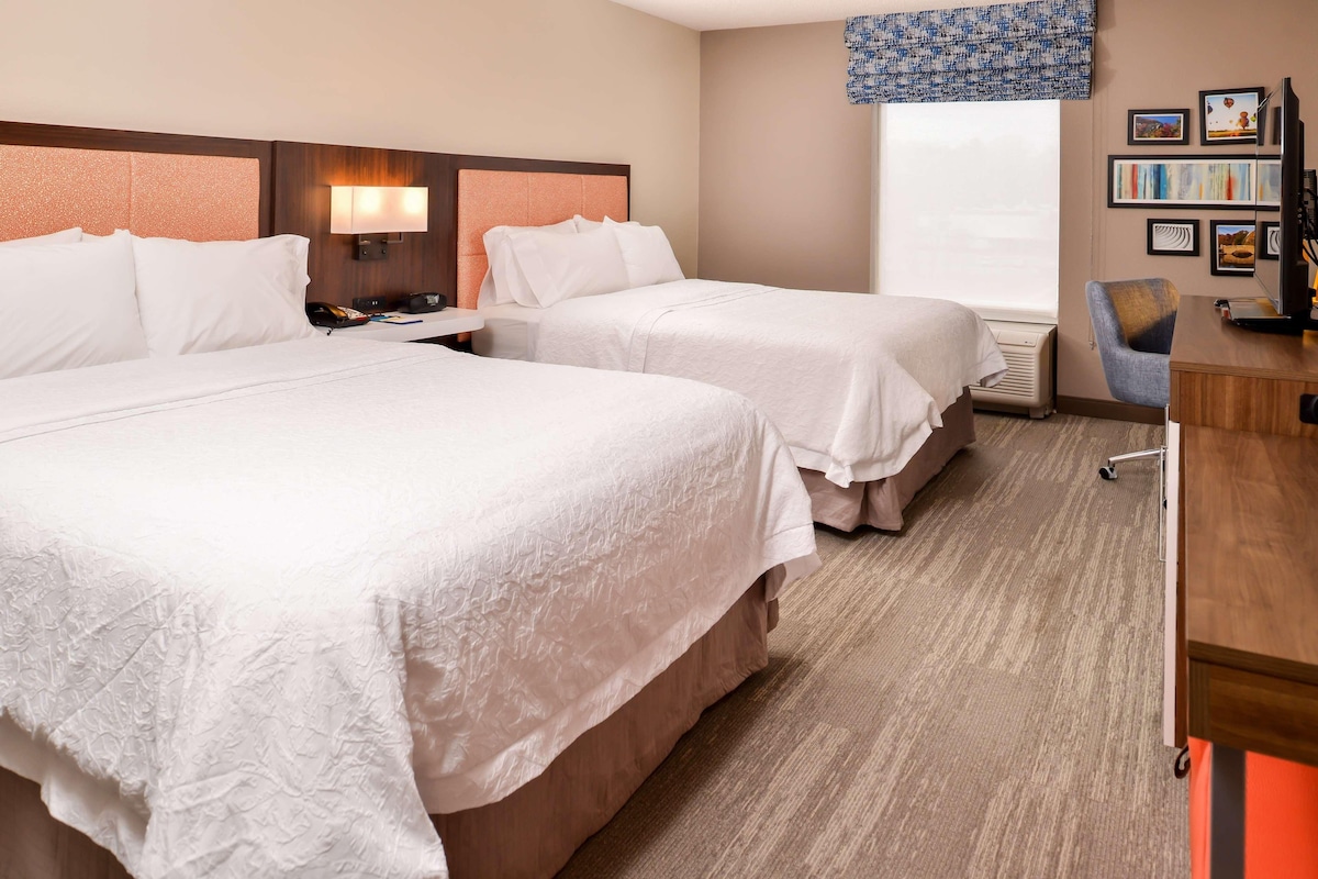 A hotel room with two beds, a desk, a chair, a TV, and a window with a view.