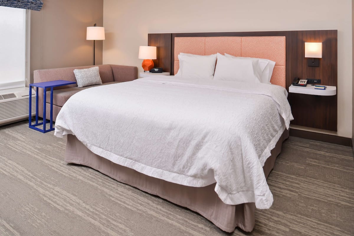 A hotel room with a large bed, a nightstand with a lamp, a small table, and a sofa.