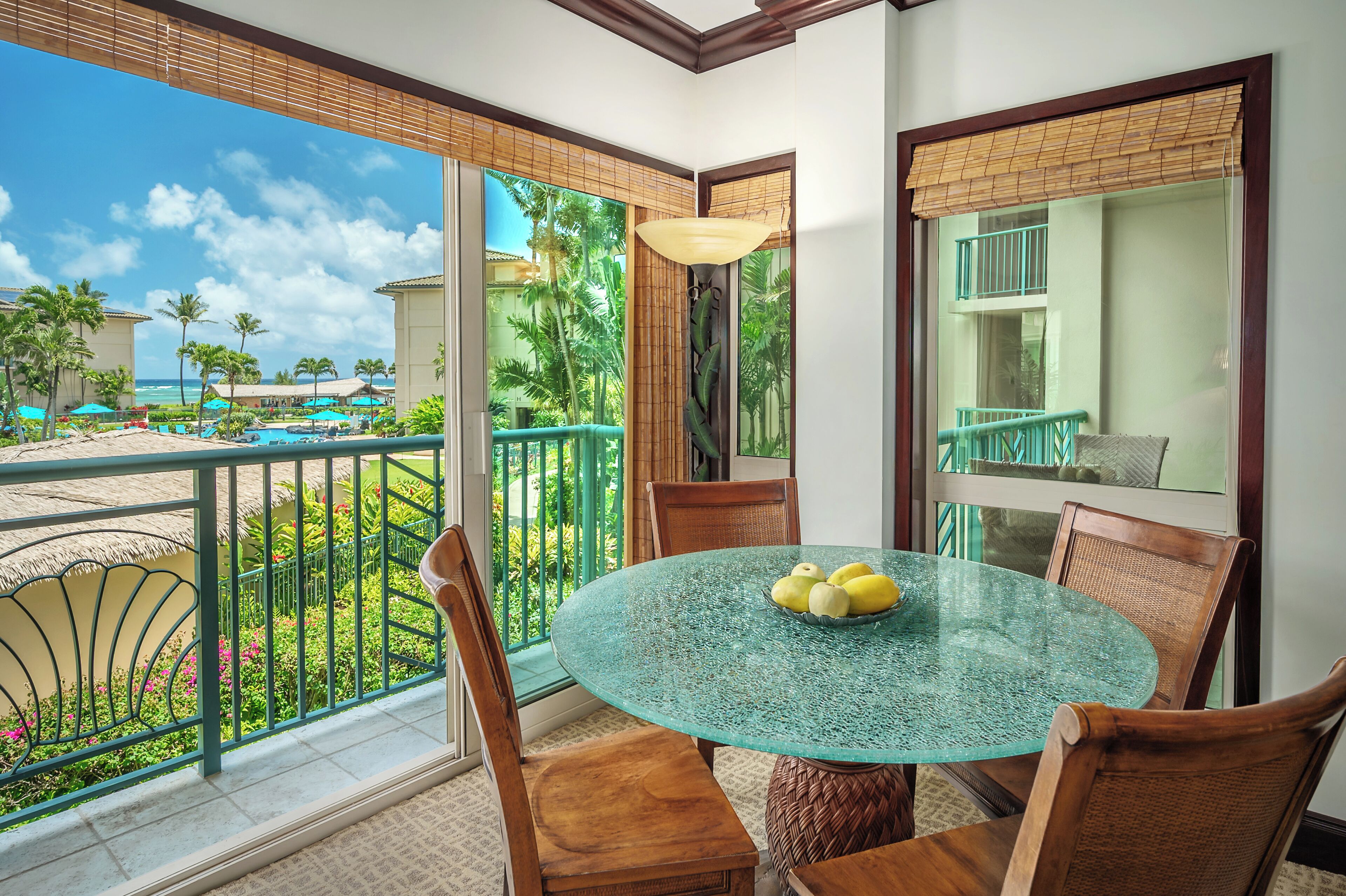 A glass table with chairs in a room with a balcony overlooking a pool and palm trees.