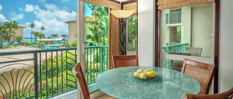 A glass table with chairs in a room with a balcony overlooking a pool and palm trees.