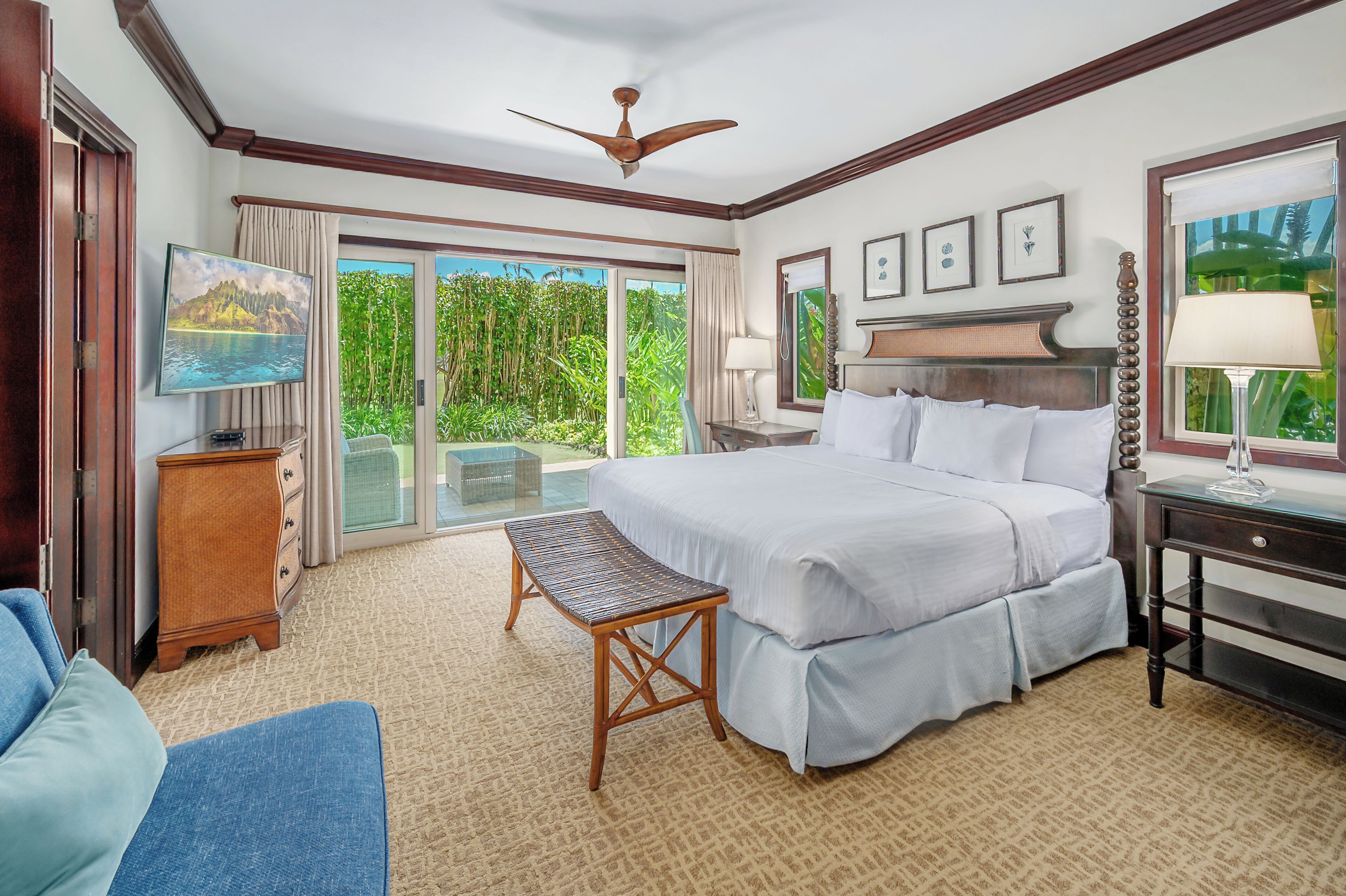 A spacious hotel room with a large bed, a desk, a chair, and a view of lush greenery through the sliding glass doors.
