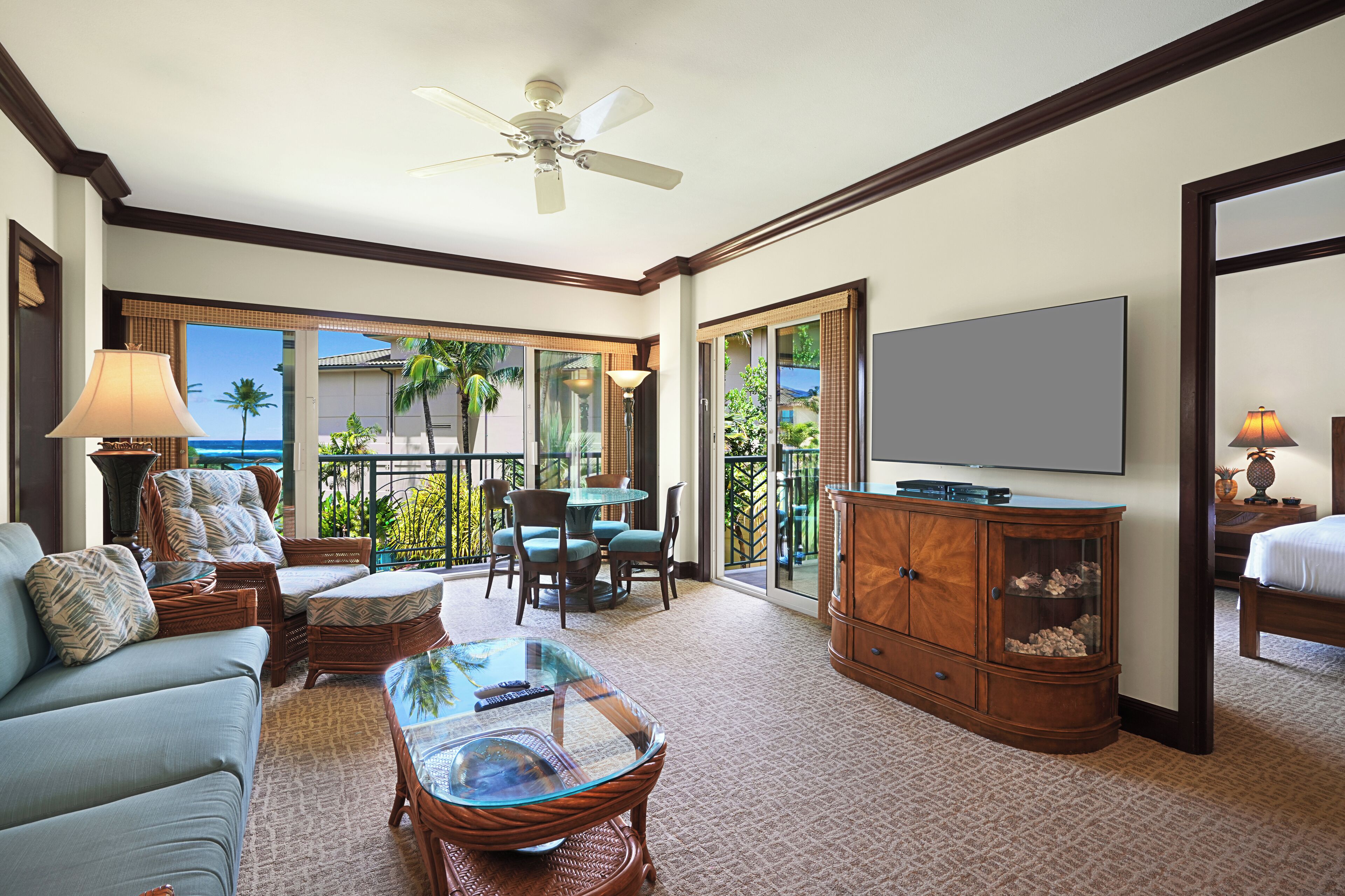 A hotel room with a sofa, chairs, a glass coffee table, a TV, a cabinet, and a balcony with a view of palm trees and the ocean.