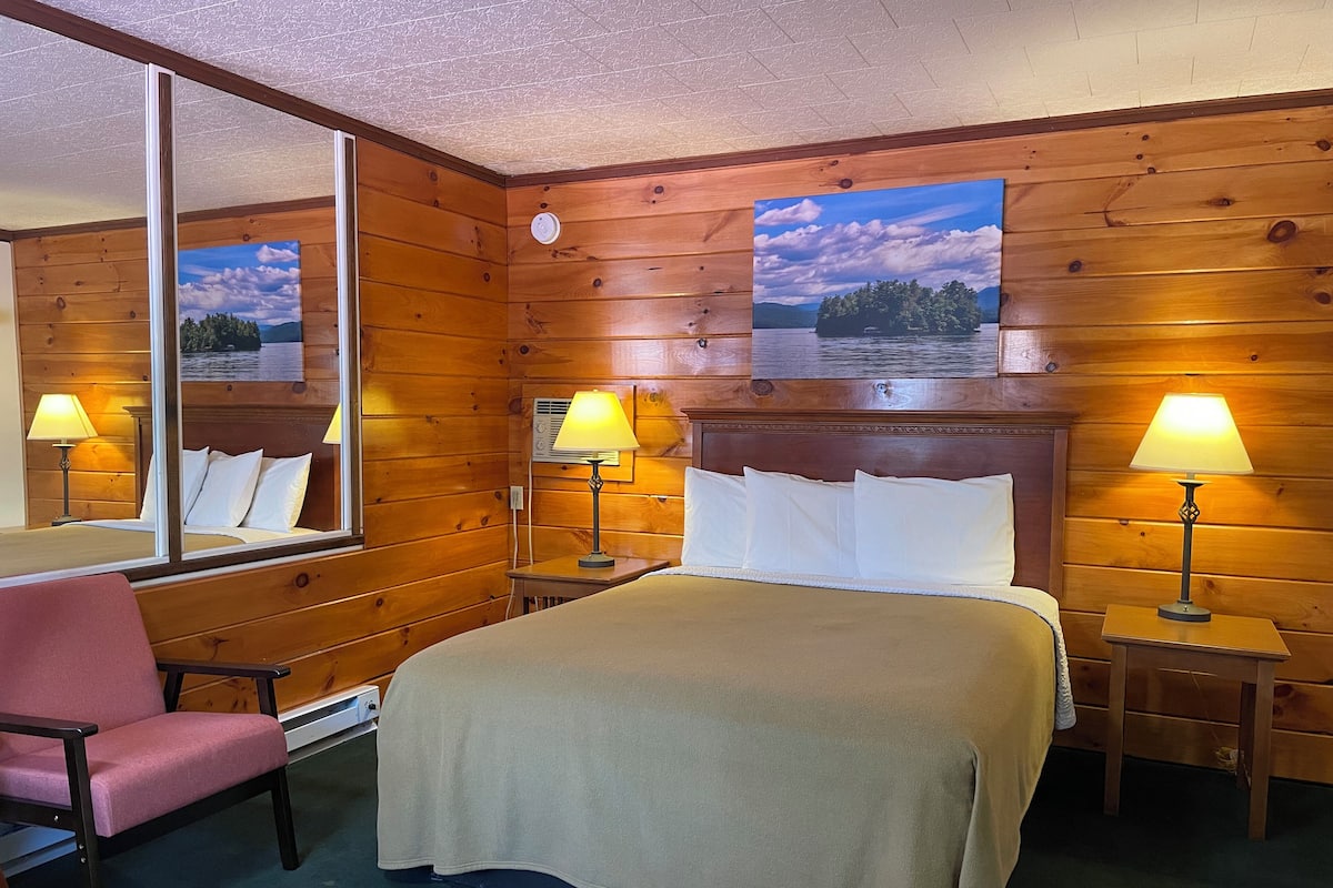A wooden-paneled room with a bed, two bedside tables with lamps, a chair, and a large mirror.