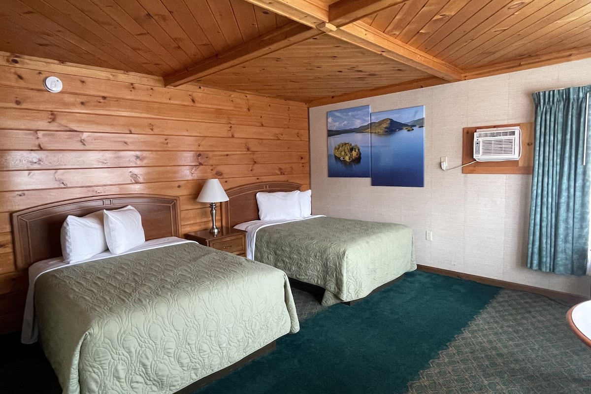 A room with two beds, wooden walls, a ceiling fan, and a window with curtains.