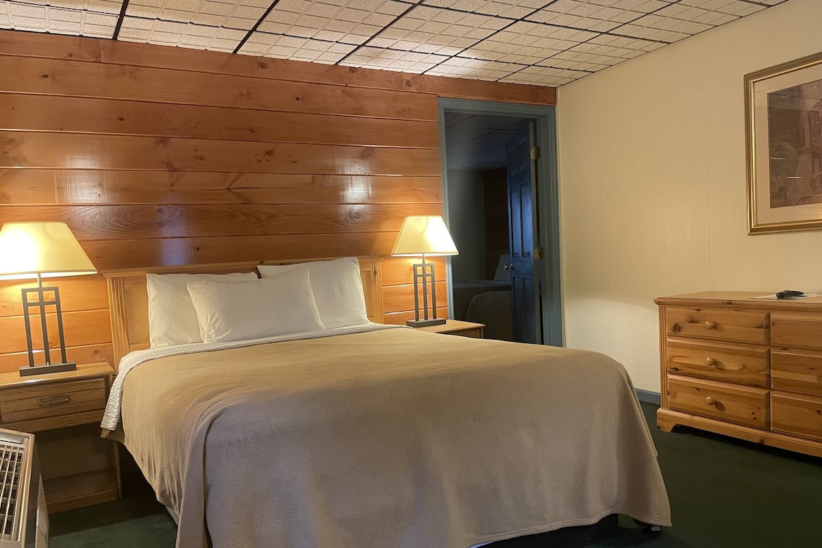A bedroom with a wooden headboard, a bed with white linens, two bedside lamps, a dresser, and a door leading to another room.