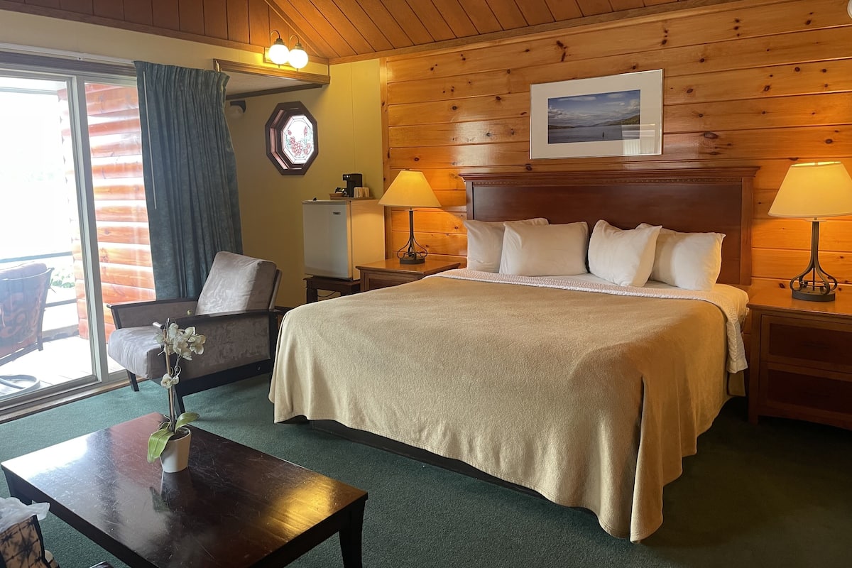 A cozy hotel room with a large bed, two bedside lamps, a small table with a vase of flowers, a chair, a desk, and a window with curtains.