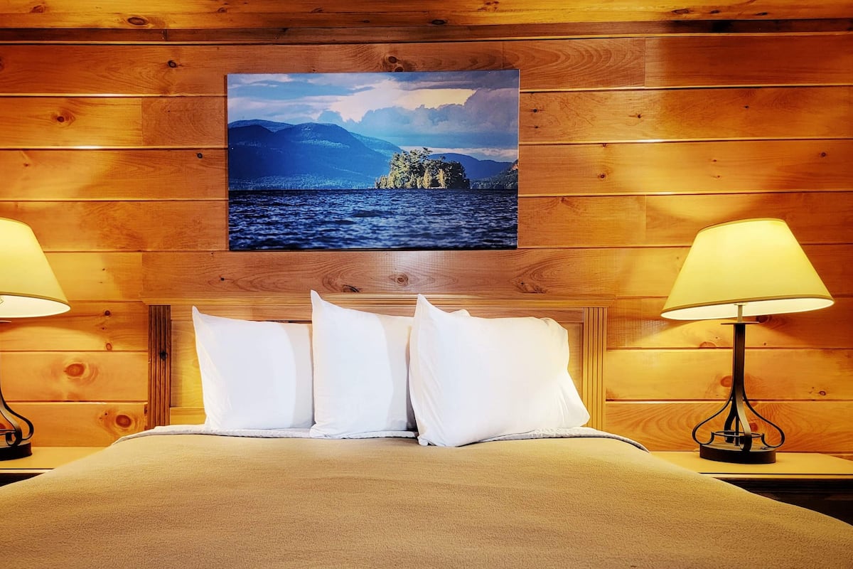 A wooden cabin room with a bed, two bedside lamps, and a scenic painting on the wall.