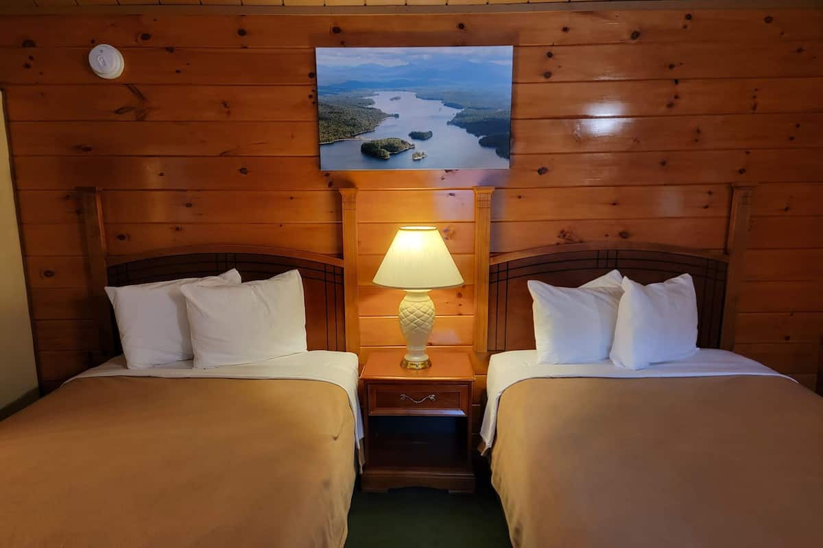 A hotel room with two beds, a nightstand with a lamp, and a painting of a landscape on the wall.