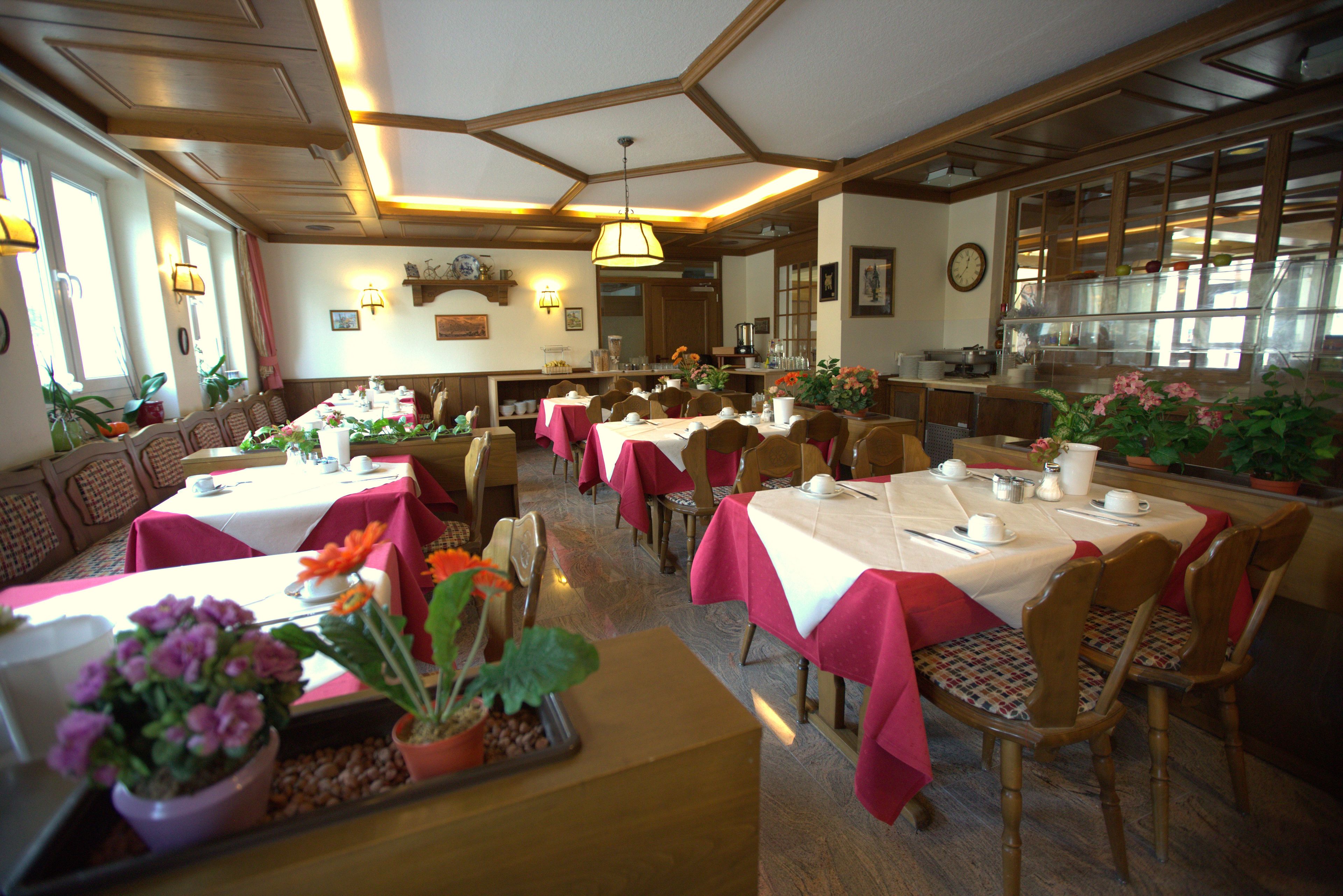 A restaurant with multiple tables and chairs, some with red tablecloths. There are potted plants on the tables and a clock on the wall.