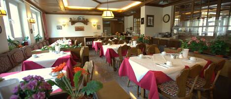 A restaurant with multiple tables and chairs, some with red tablecloths. There are potted plants on the tables and a clock on the wall.