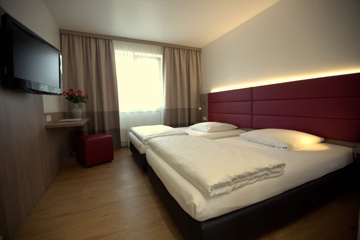 Habitación de hotel con dos camas, un televisor, un pequeño taburete rojo y un florero con flores.