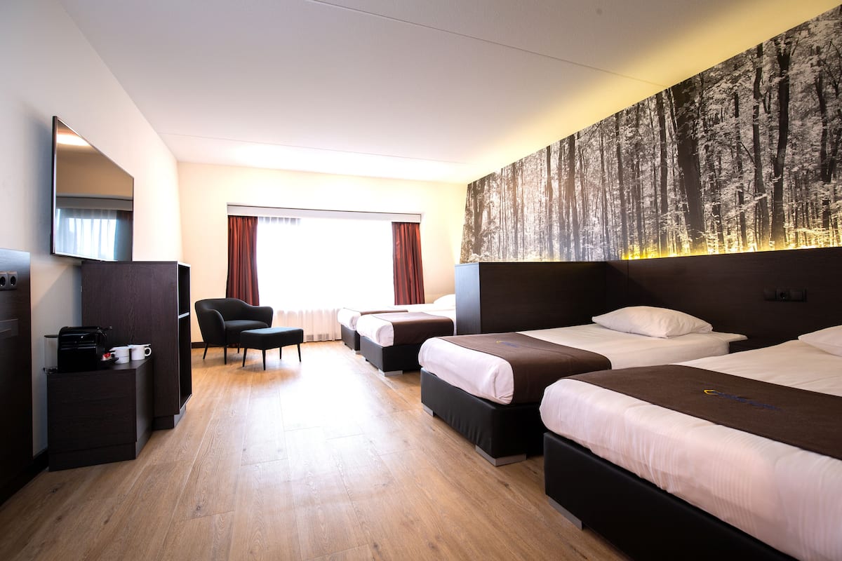 Ein Hotelzimmer mit drei Betten, einem Schreibtisch mit Computer, einem Sessel und einem großen, an der Wand befestigten Fernseher.