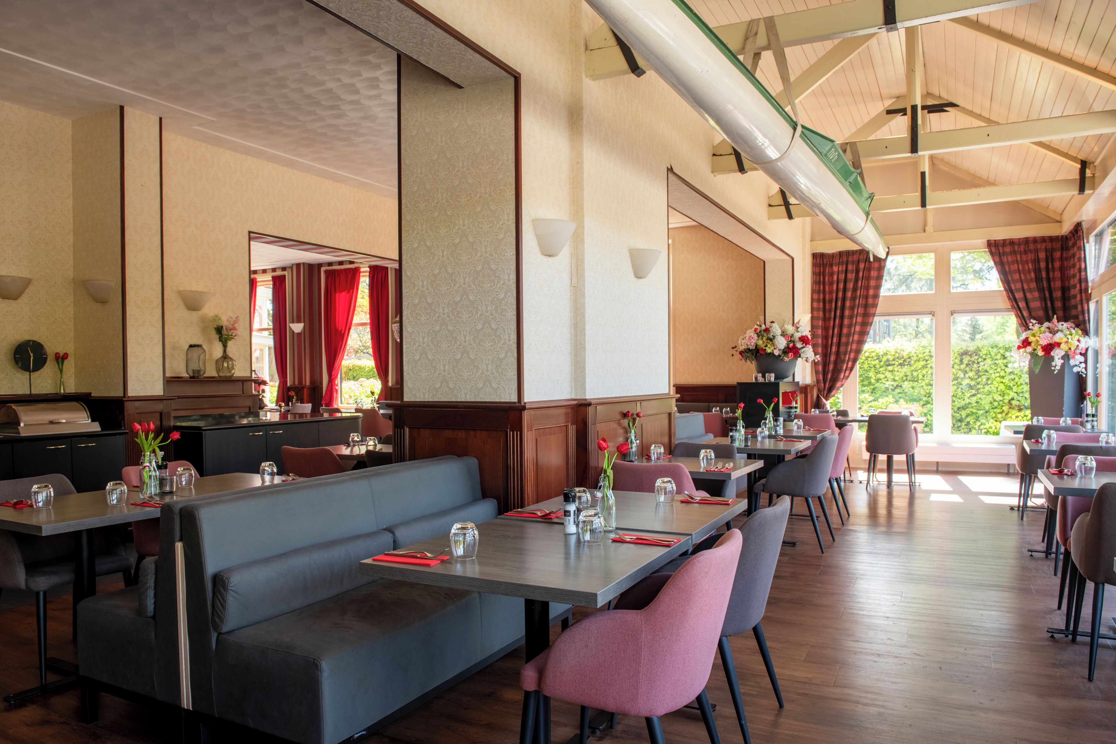 A spacious restaurant with high ceilings, wooden beams, and large windows. Tables are set with red napkins and glassware.