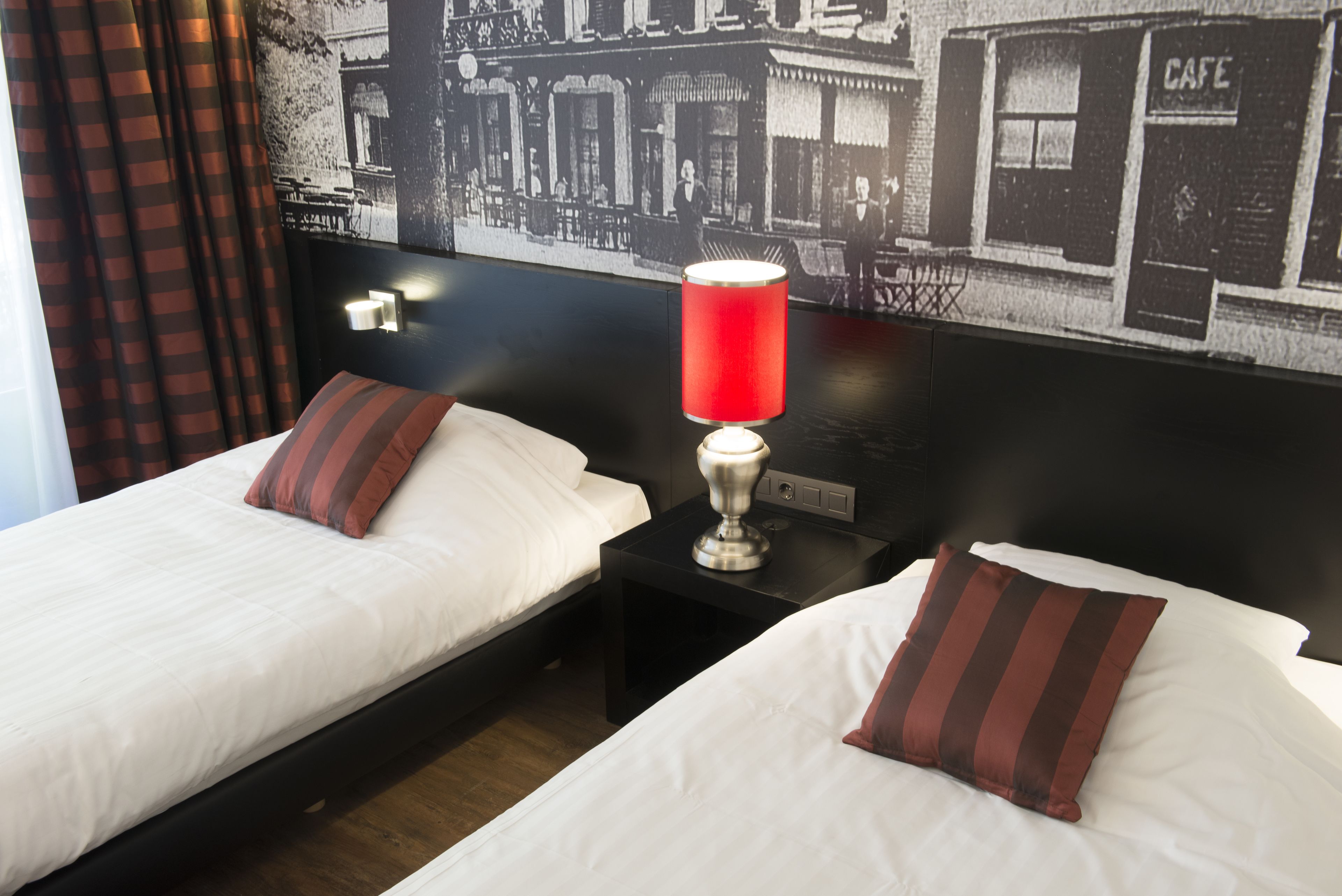Quarto de hotel com duas camas, cada uma com lençóis brancos e almofadas listradas, uma luminária de mesa vermelha e uma mesa de cabeceira preta. Uma fotografia em preto e branco de um café está na parede atrás das camas.
