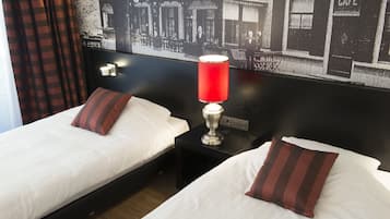 A hotel room with two beds, each with white linens and striped pillows, a red table lamp, and a black nightstand. A black and white photo of a cafe is on the wall behind the beds.