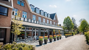 A brick building with large windows and a covered walkway. There are potted plants and bicycles outside. - Bastion Hotel Apeldoorn Het Loo (Apeldoorn)