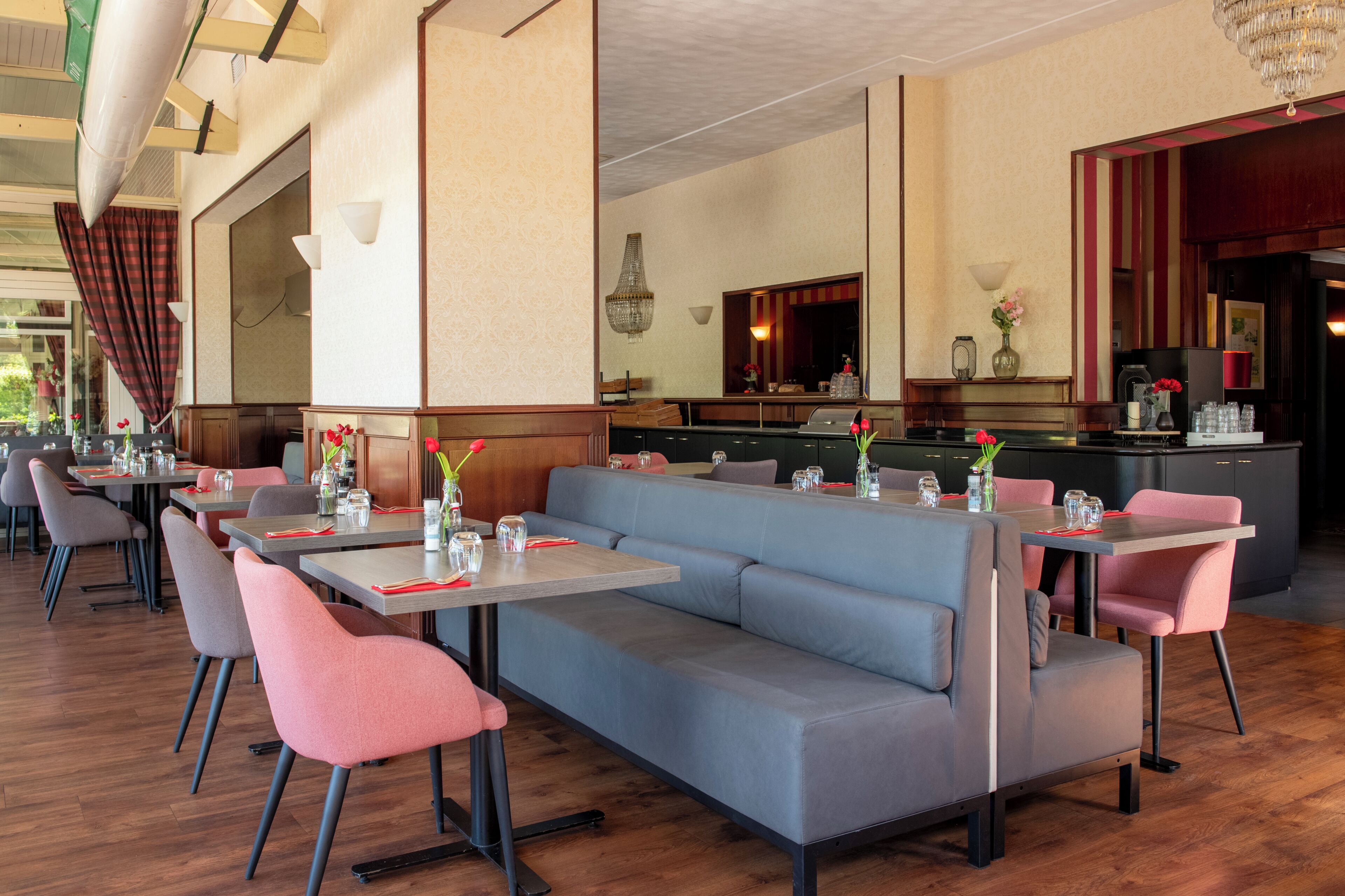 Un ristorante con un lungo divano grigio, sedie rosa e tavoli apparecchiati con tovaglioli rossi e bicchieri.