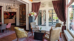 A spacious living area with a large window, a chandelier, and a fireplace. - Bastion Hotel Apeldoorn Het Loo (Apeldoorn)