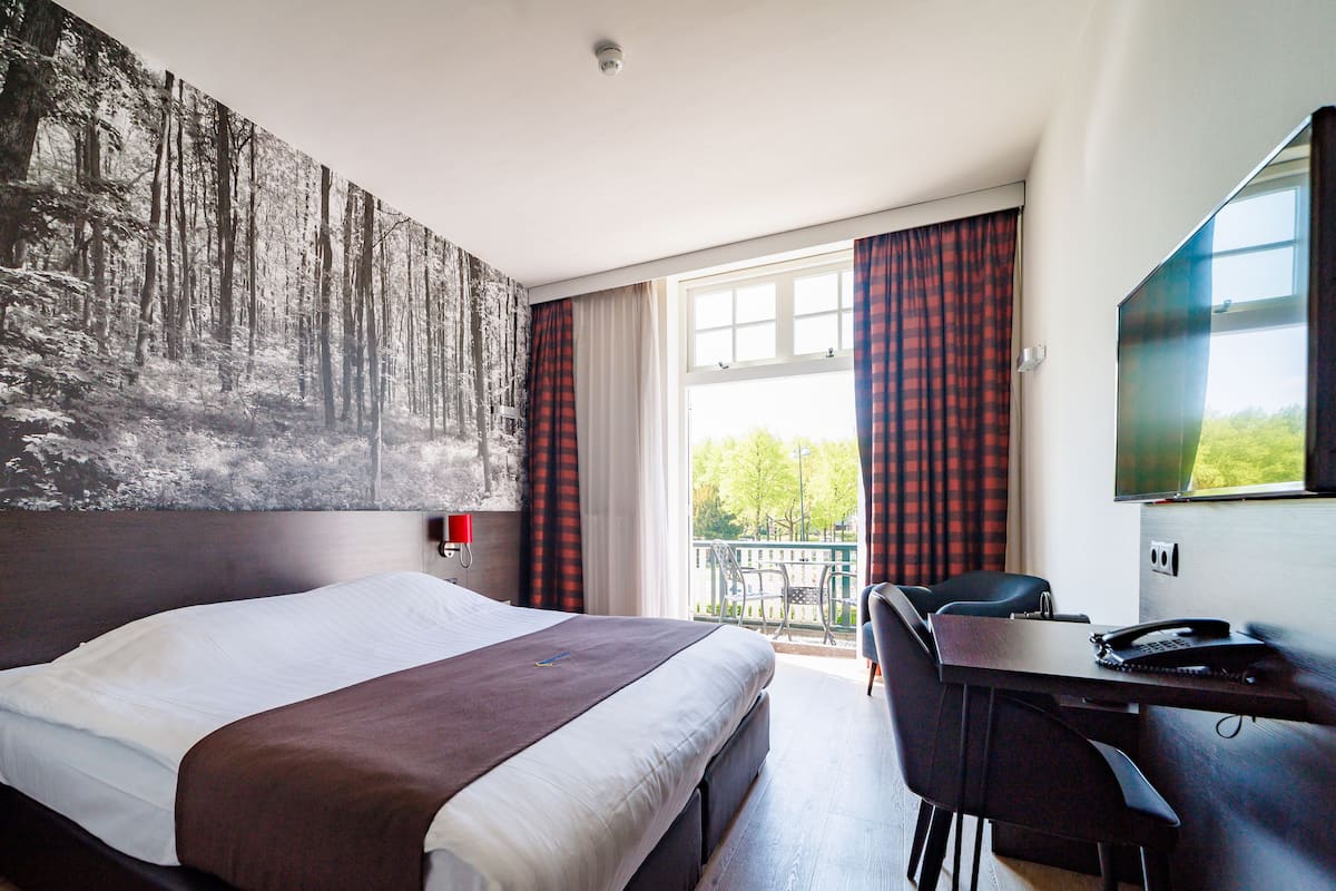 Ein Hotelzimmer mit einem großen Bett, einem Schreibtisch mit Stuhl, einem Fernseher und einem Balkon mit roten Vorhängen.