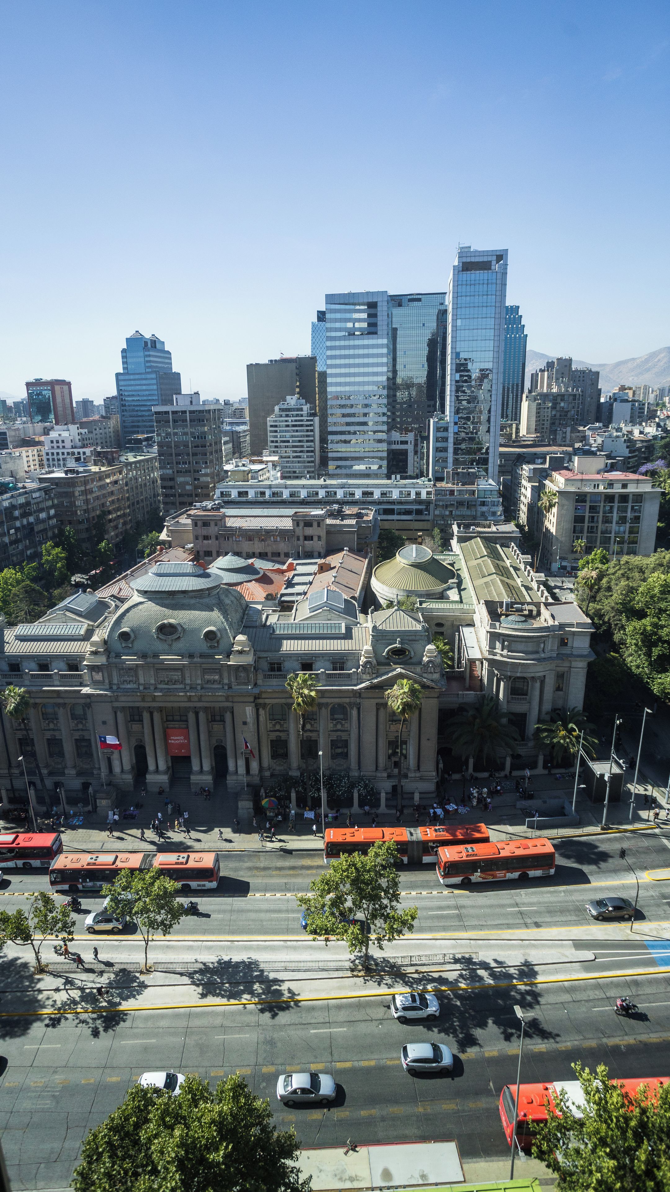 A cityscape with modern buildings, a central plaza with a dome structure, and a busy street with buses and cars.