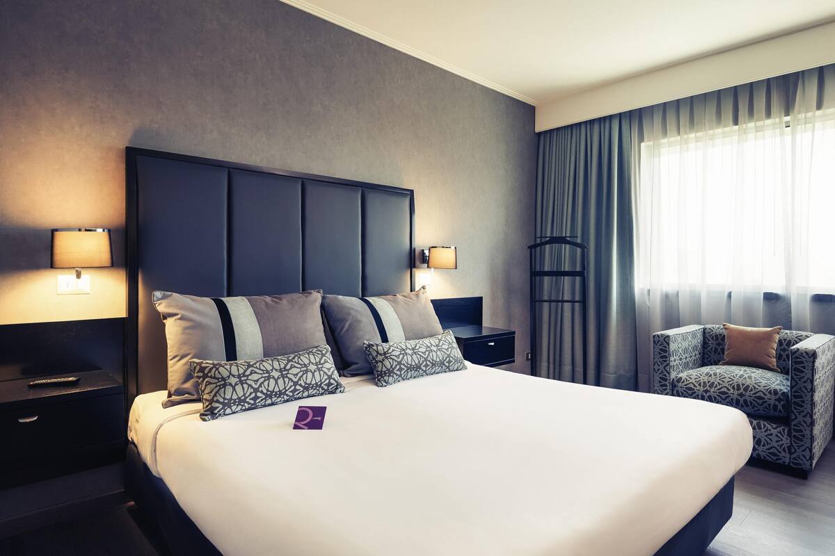 Habitación de hotel con una cama grande, lámparas de noche, una silla, una mesita de noche y una ventana con cortinas.