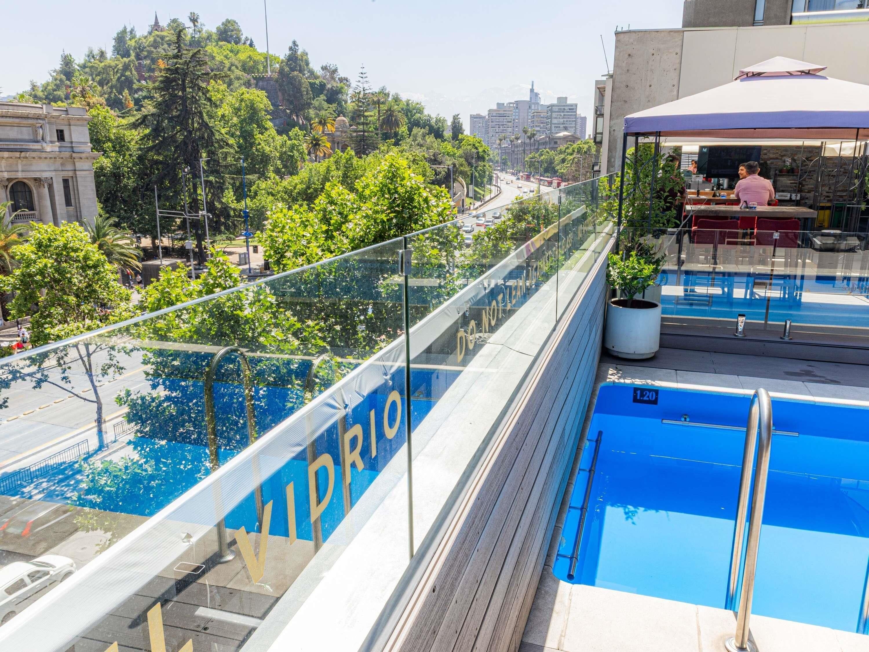 Una piscina en la azotea con barandilla de cristal, una zona de bar y vistas a la ciudad.