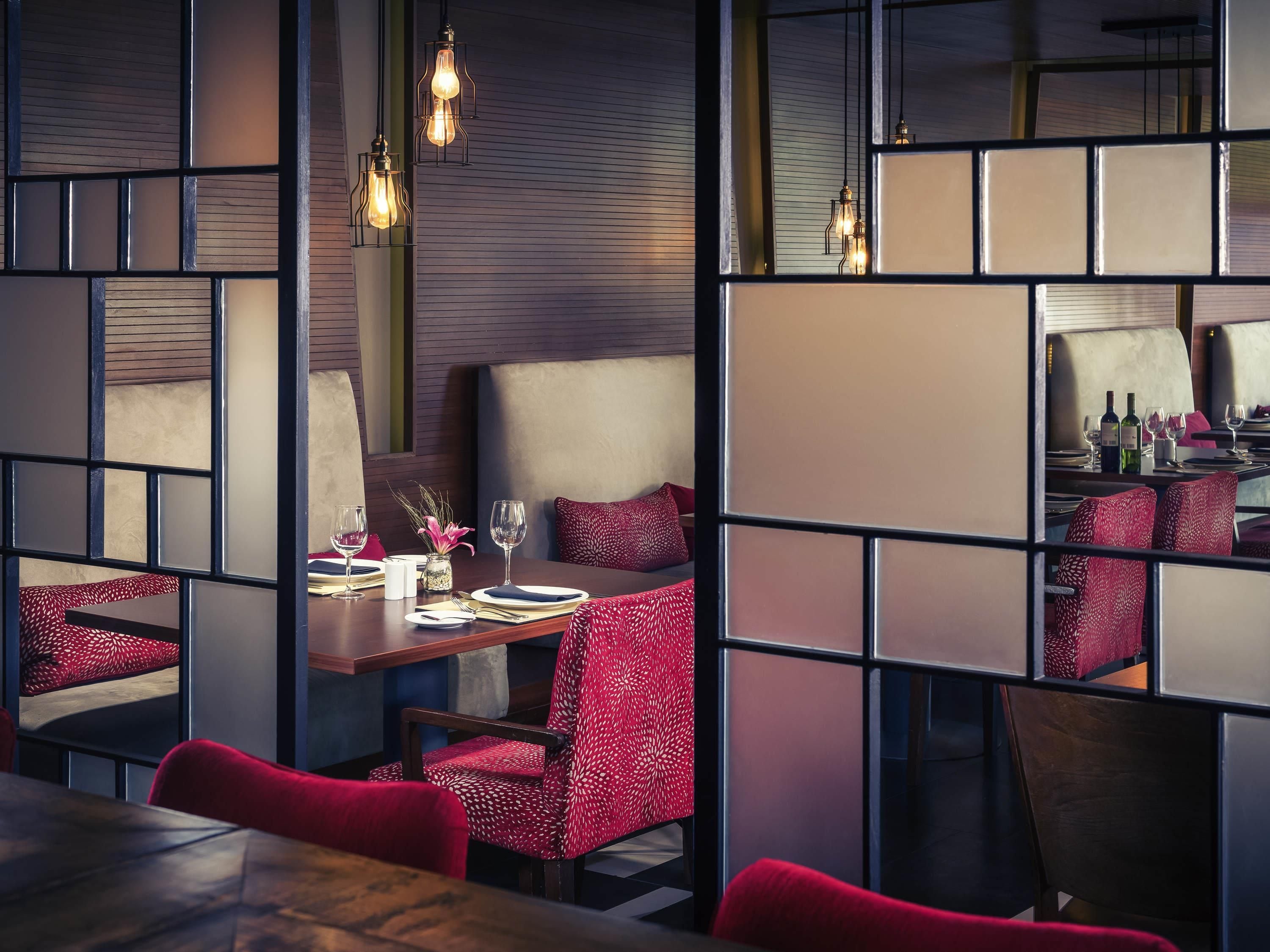A restaurant with a modern interior, featuring a wooden table set for two, red upholstered chairs, and hanging pendant lights.