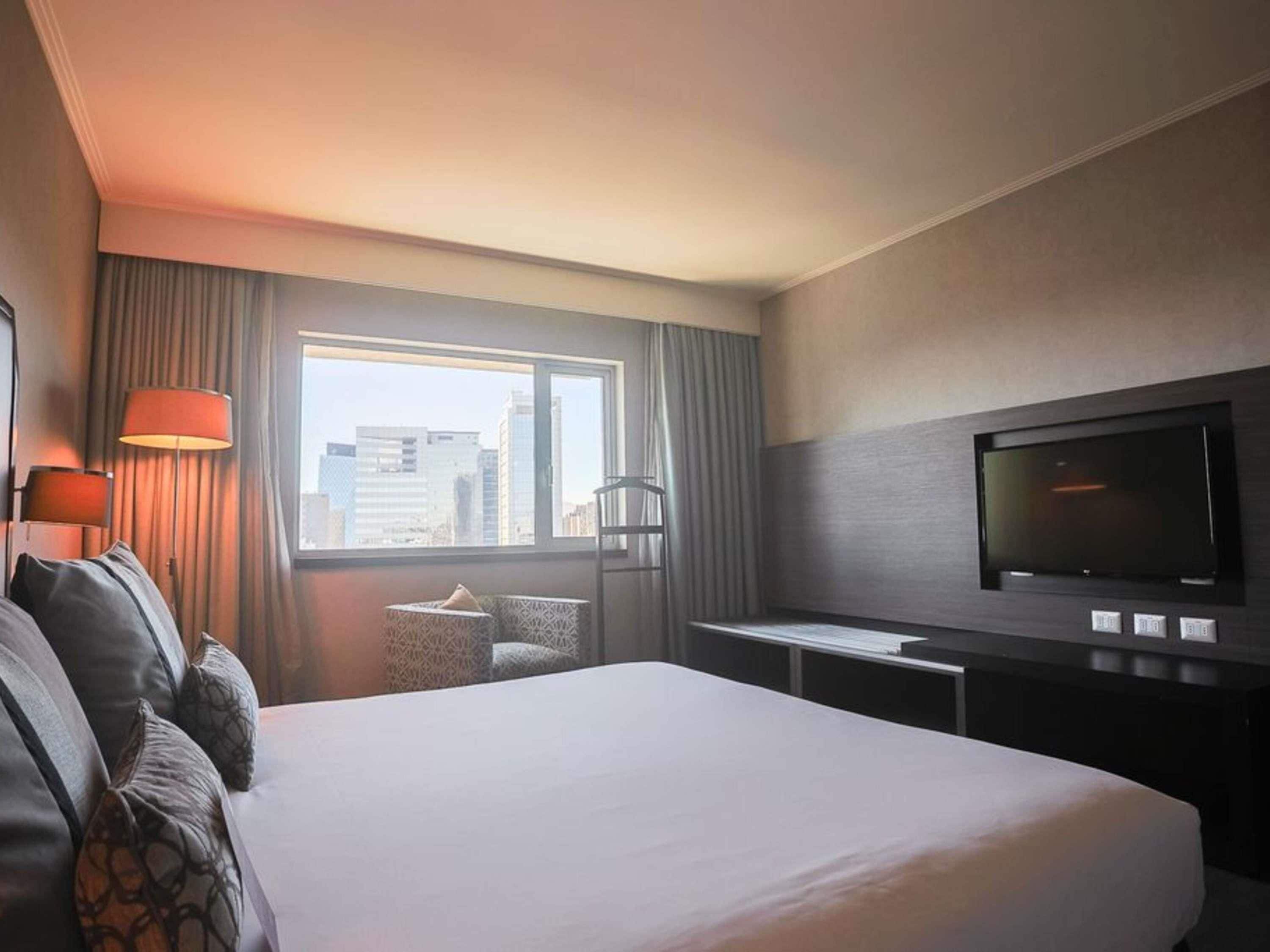 Quarto de hotel com uma cama grande, televisão na parede, janela com vista para a cidade e mesa de trabalho com abajur.