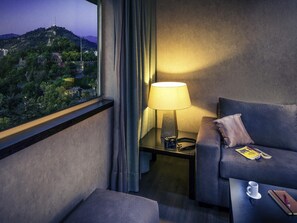 A hotel room with a view of a hilltop, a sofa, a bedside table with a lamp, and a small table with a cup and a book. - Mercure Santiago Centro (Santiago)