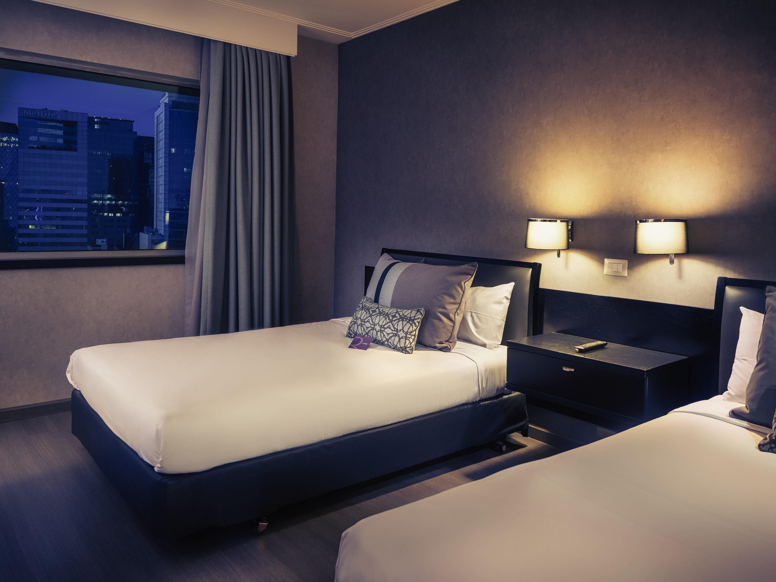 Ein Hotelzimmer mit zwei Betten, einem Nachttisch, einer Lampe und Blick auf die Stadtlandschaft.