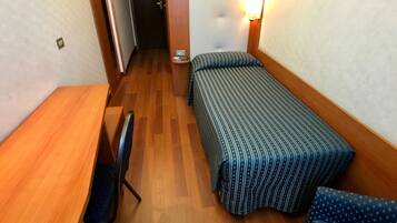 Habitación de hotel pequeña con un escritorio de madera, una cama individual con colcha estampada, una silla y una lámpara fijada a la pared.