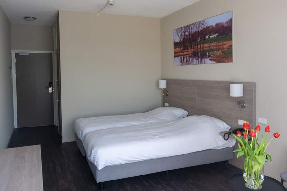 Una habitación de hotel con cama, escritorio, silla, un jarrón con flores y un cuadro en la pared.