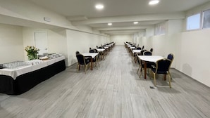 Een vergaderzaal met rijen tafels en stoelen, een buffet aan de linkerkant, en een deur aan de linkerzijde.