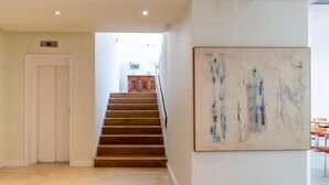 Een modern interieur met een trap naar een kamer met uitzicht, een groot abstract schilderij aan de wand, en een deur met een liftknop.
