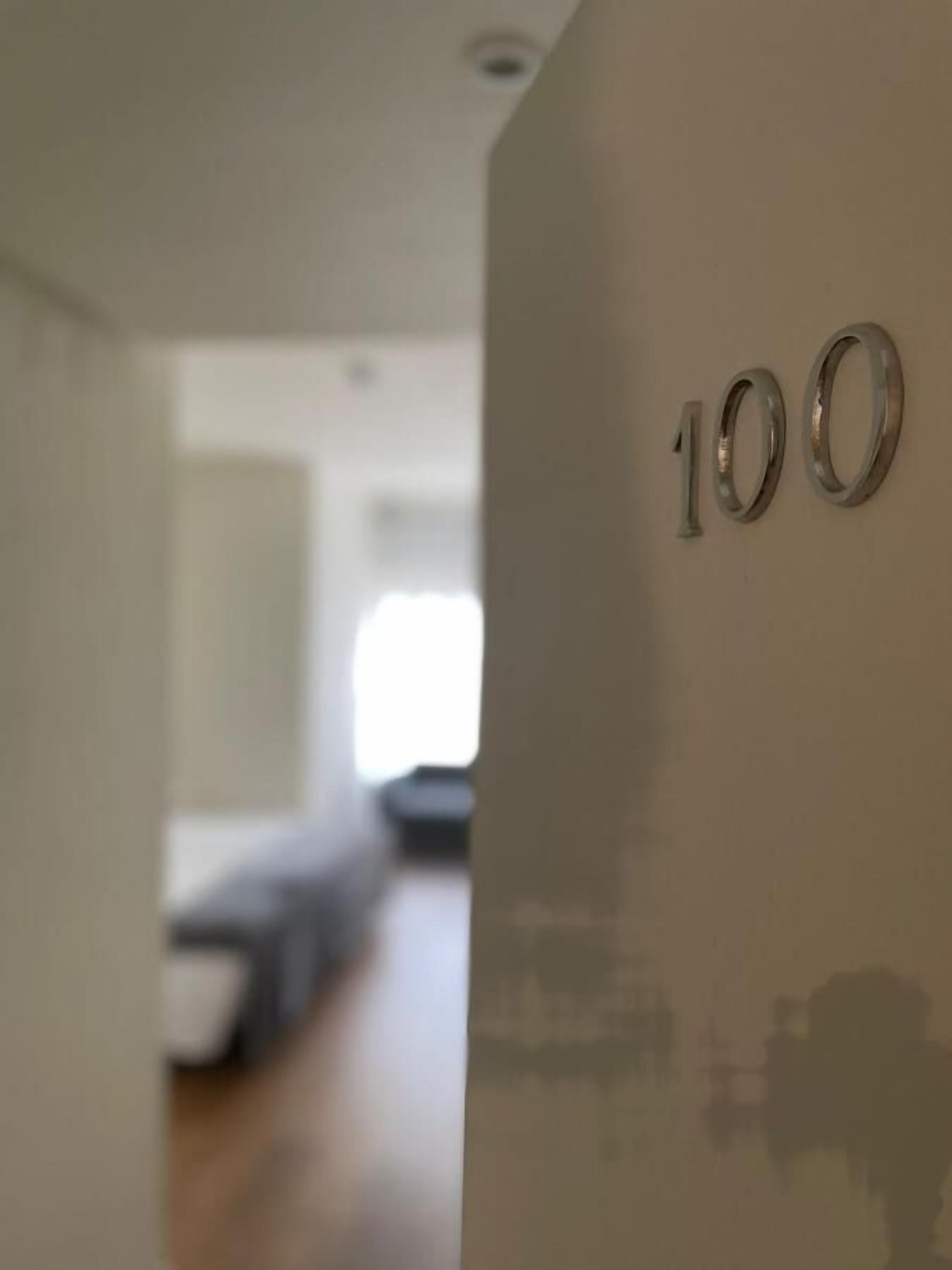 A hallway with a number '100' on the wall, leading to a room with a bed and a window.