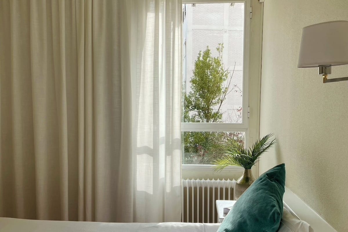 Een hotelkamer met een bed, een raam met gordijnen, een radiator en een wandlamp.