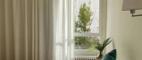 A hotel room with a bed, a window with curtains, a radiator, and a wall-mounted lamp.