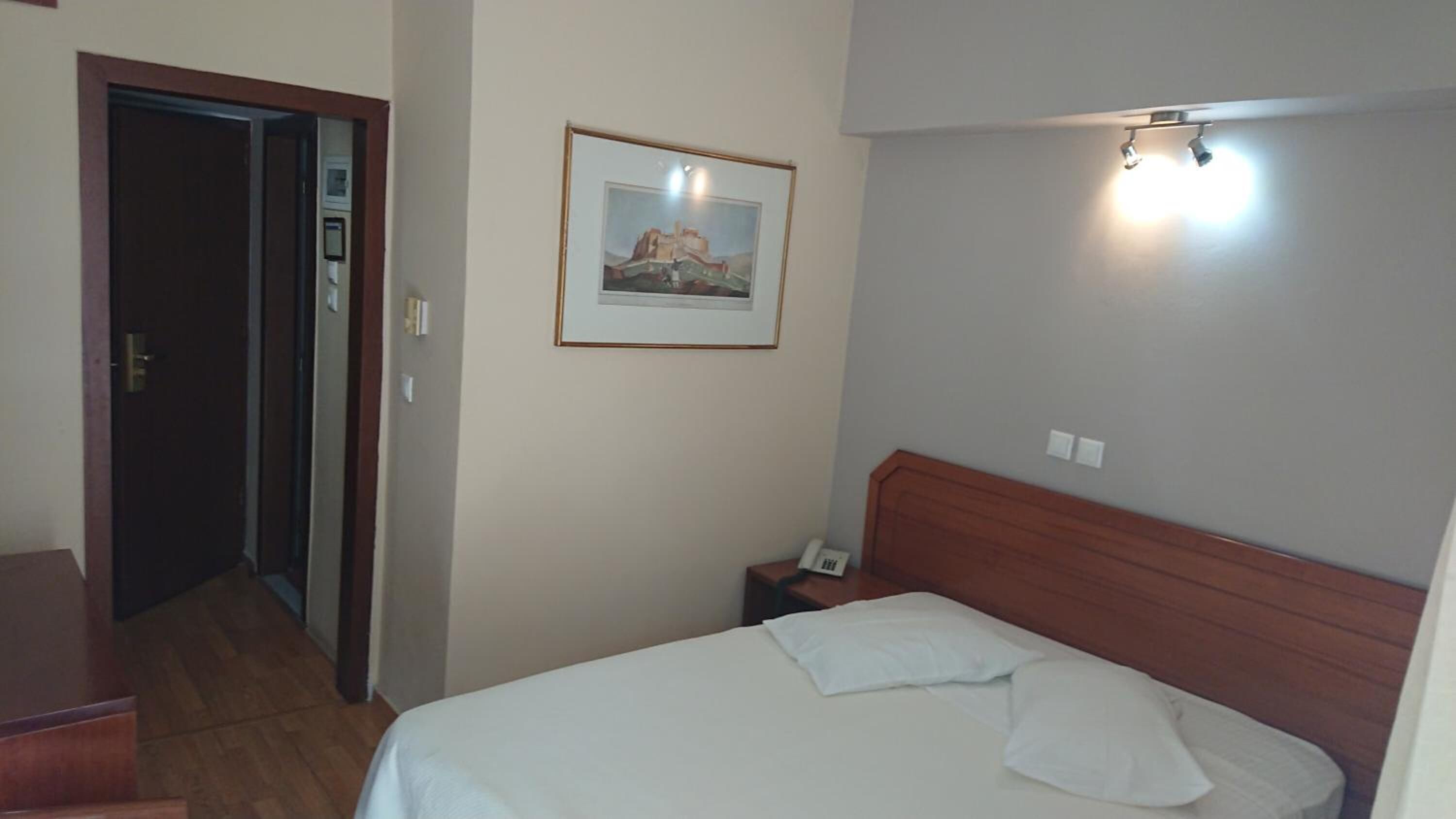 A hotel room with a bed, a nightstand with a phone, a framed picture on the wall, and a door leading to another room.