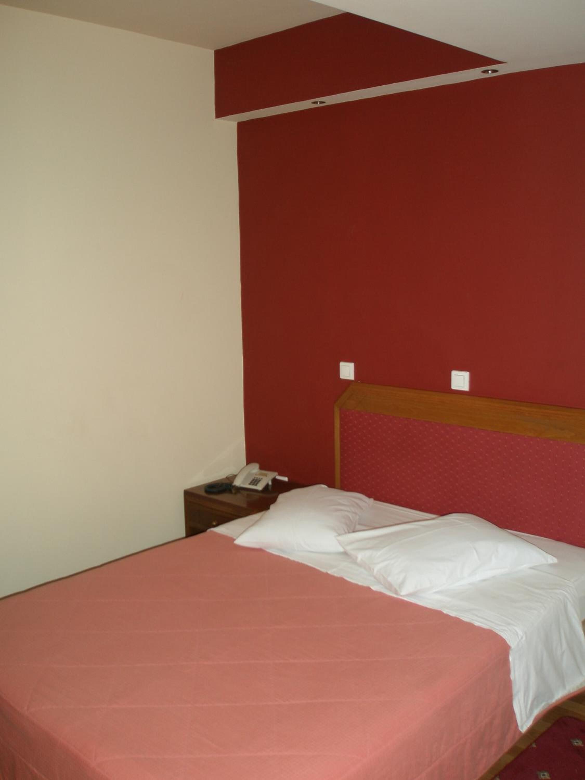 A single bed with a wooden headboard, a red wall, and a small bedside table with a phone.